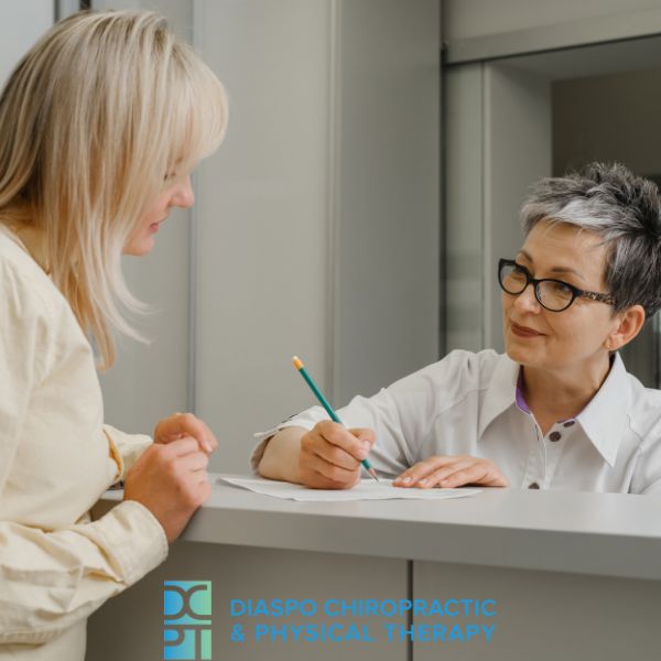 A woman stands at a reception desk while another woman, seated, writes on a notepad. The logo for Diaspo Chiropractic & Physical Therapy is visible. A woman stands at a reception desk while another woman, seated, writes on a notepad. The logo for Diaspo Chiropractic & Physical Therapy is visible.
